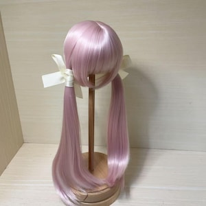 May include: A light pink wig with long pigtails, secured with white ribbons. The wig is displayed on a wooden stand, showcasing its smooth, shiny texture and style. The wig's color is a soft, pastel pink.