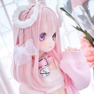 May include: A pink doll with pink hair, purple eyes, and white horns. The doll wears a pink outfit with a collar and a silver bell. The doll's hair is styled with braids and a bow.