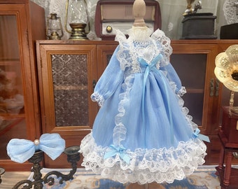 Puede incluir: Un vestido de muñeca azul claro con mangas largas, ribete de encaje blanco y un detalle de encaje en forma de corazón en el corpiño. También hay un lazo azul a juego y un lazo para el pelo.