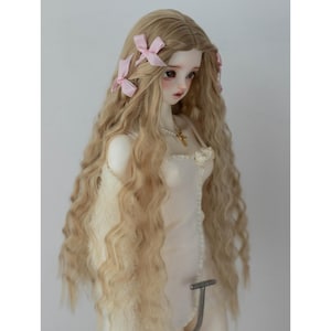 May include: A porcelain doll with long, wavy blonde hair decorated with pink bows. The doll is dressed in a cream-coloured dress and a delicate necklace with a gold pendant. The doll is a collectible item.