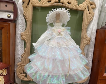 Puede incluir: Un vestido blanco con una falda de capas y tela iridiscente. El vestido tiene un corpiño de encaje blanco con un acento de flor rosa. El vestido está en un maniquí que lleva un sombrero blanco con ribete de encaje.