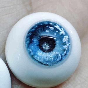 May include: A close-up of a blue glass eye with a white sclera. The eye has a realistic appearance with a dark pupil and a blue iris with white flecks.