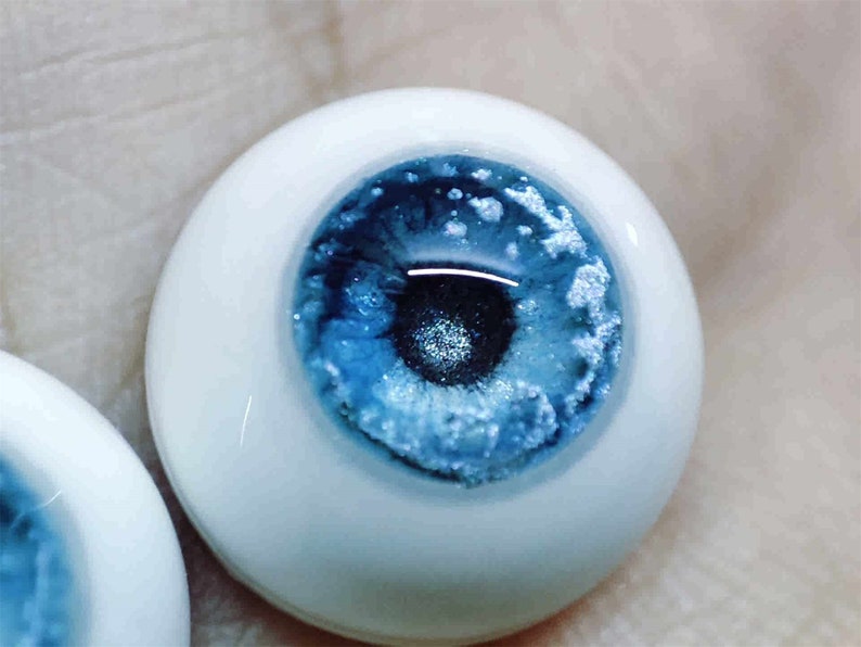 May include: A close-up of a blue glass eye with a white sclera. The eye has a realistic appearance with a dark pupil and a blue iris with white flecks.