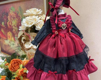 Puede incluir: Un vestido de muñeca rojo y negro con ribete de encaje y un lazo rojo. El vestido tiene un broche en forma de corazón con pedrería. La muñeca lleva un sombrero rojo a juego con un lazo negro.