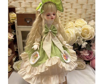 May include: A porcelain doll with long blonde hair and a green and cream-colored dress. The doll wears a matching hat and has a large bow at the waist. The dress features floral embroidery and ruffled trim. The doll is posed on a small wooden stool.