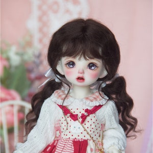May include: A porcelain doll with dark brown pigtails, wearing a white knitted sweater and a red and white polka dot dress. The doll has large, purple eyes and rosy cheeks. A small gold chain purse hangs from the doll's shoulder.