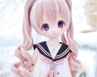 May include: A doll with pink pigtails, dressed in a white sailor-style dress with a black collar and pink details. The doll has large eyes with purple and pink tones, and rosy cheeks. A small gold crescent moon charm is on the dress.