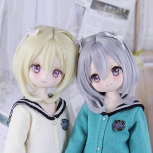 May include: Two anime-style dolls with large, expressive eyes and soft, blushed cheeks. One doll has blonde hair, the other has gray hair, both styled with a bow. They wear matching school uniform-style sweaters with a crest.