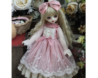 Puede incluir: Una muñeca rosa y blanca con un vestido de encaje rosa y blanco con un lazo en el pelo. La muñeca tiene el pelo rubio y los ojos azules.