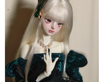 May include: A white porcelain doll with long, straight, white hair. The doll is wearing a green velvet dress with a pearl necklace and a peacock feather hair accessory.