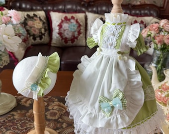 Puede incluir: Un vestido verde claro y blanco con un sombrero a juego, exhibidos en soportes de madera. El vestido presenta un cuello, mangas y delantal con volantes, con un lazo a cuadros. El sombrero tiene un borde con volantes y ribete de encaje.