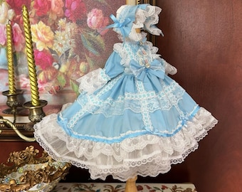 Puede incluir: Un vestido de muñeca azul claro con ribete de encaje blanco y un sombrero a juego. El vestido tiene un lazo en la cintura y una falda de capas.