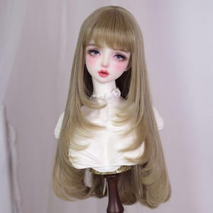 May include: A doll head with long, layered, light brown hair and a fringe. The doll has delicate facial features, including pink blush, blue eyes, and red lips. It is wearing a white top with a floral necklace.