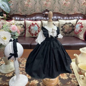 May include: A black and white doll's dress with a white ruffled collar and a silver cross pendant. The dress has a black bow at the waist and a black skirt with white stitching.