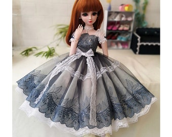May include: A doll wearing a black and white tulle dress with blue floral embroidery. The dress has a white lace trim and a white bow at the waist.