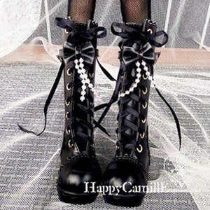 Op de afbeelding: Zwarte leren laarzen met zwarte veters en een zwarte strik bovenop elke laars. De laarzen hebben een witte parelketting die aan de strik hangt.