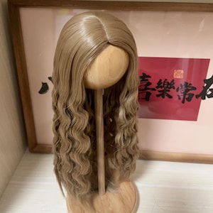 May include: A light brown wig with long, wavy curls is displayed on a wooden stand. The wig has a natural-looking part and is styled with loose waves. The stand is set against a background with framed artwork.