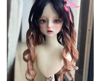 May include: A doll wig with long, curly hair. The wig is a dark brown color at the roots and fades to a light brown color at the ends. The wig is styled with a side part and has a small pink bow with a rhinestone accent.