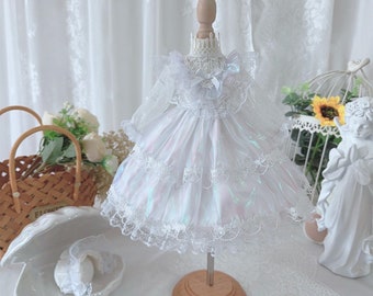 Puede incluir: Un vestido blanco de tul iridiscente de varias capas con ribete de encaje, exhibido en un maniquí de madera. El vestido tiene un escote con volantes y un lazo en el centro.