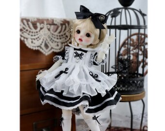 May include: A white and black doll wearing a white dress with black trim and a black bow in her hair.