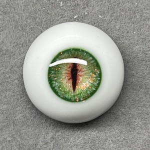 Puede incluir: Primer plano de un ojo de vidrio verde y marrón con purpurina. El ojo está fijado en una base de plástico blanco.