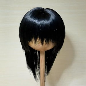 May include: A black wig with a blunt fringe and long, layered sides is displayed on a wooden wig stand. The wig has a shiny, smooth texture, and the fringe is cut straight across the forehead.