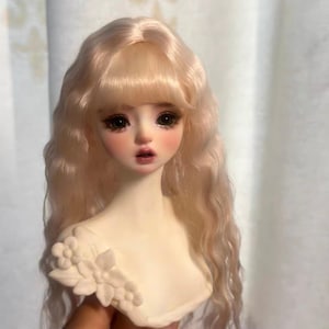 May include: A doll head with long, wavy, light blonde hair and bangs. The doll has detailed facial features, including large, expressive eyes, rosy cheeks, and a slightly open mouth. A floral embellishment adorns the shoulder.
