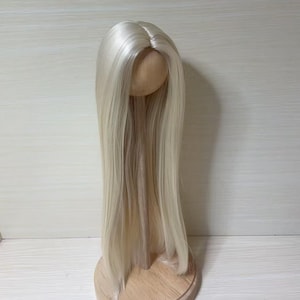 May include: A doll wig with long, straight, platinum blonde hair. The wig is mounted on a wooden head form and displayed against a light background. The hair is parted in the middle and falls in a smooth, flowing style.