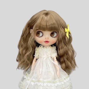 May include: A Blythe doll with long, wavy brown hair and large, brown eyes. The doll is dressed in a white lace dress, with a yellow bow in her hair. The doll has a pale complexion and rosy cheeks.