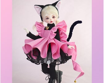 May include: A pink and black doll with a cat costume. The doll has blonde hair, blue eyes, and is wearing a pink dress with black trim. The doll has a black cat tail and a white cat ear headband.