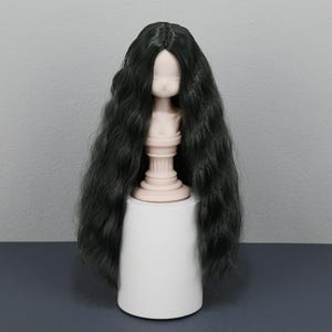 May include: A doll head with long, wavy, dark green hair. The doll head is mounted on a light pink pedestal, which is placed on a white cylindrical base. The background is a solid grey.