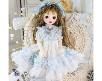 Puede incluir: Una muñeca de porcelana con un vestido blanco con ribetes de encaje azul y una diadema azul. La muñeca tiene el pelo largo y castaño y los ojos azules.