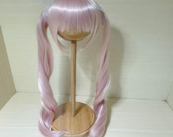 Puede incluir: Una peluca rosa claro con largas coletas fluidas. La peluca se exhibe en un soporte de madera con una base redonda. El cabello tiene una ligera onda y es de un color pastel suave.