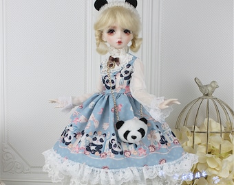 May include: A blue and white doll dress with a panda print and a white lace trim. The dress has a black chain strap with a panda-shaped purse attached. The doll is wearing a white blouse with a black bow and black panda ears.