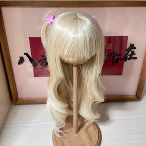 May include: A blonde wig with bangs and long, wavy layers, styled with two small pink flower hair clips. The wig is displayed on a wooden stand, with a framed artwork in the background.