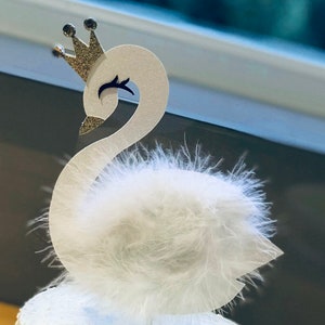 K&ouml;nnte beinhalten: Ein wei&szlig;er Schwan-Kuchenaufsatz mit einer goldenen Glitzerkrone und flauschigen Federakzenten. Der Schwan hat ein schwarzes Auge und einen goldenen Glitzerschnabel. Der Aufsatz befindet sich auf einem wei&szlig; glasierten Kuchen.