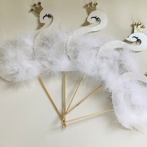 K&ouml;nnte beinhalten: Schwanf&ouml;rmige Partydekorationen mit wei&szlig;en Federn und goldenen Glitzerkronen. Die Schw&auml;ne sind an Holzst&auml;ben befestigt und erzeugen einen skurrilen und eleganten Look. Perfekt f&uuml;r eine Babyparty oder eine Geburtstagsfeier.