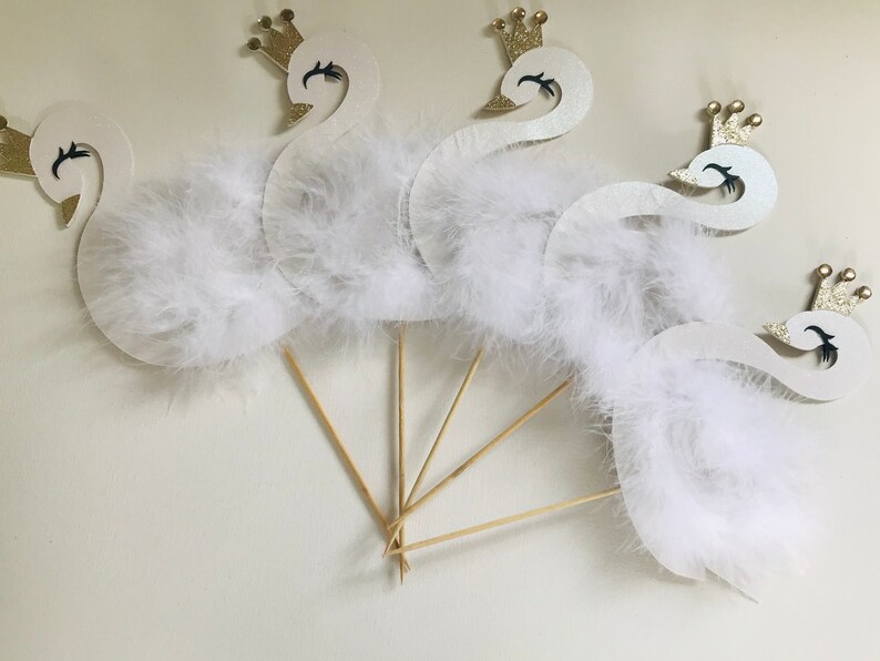 K&ouml;nnte beinhalten: Schwanf&ouml;rmige Partydekorationen mit wei&szlig;en Federn und goldenen Glitzerkronen. Die Schw&auml;ne sind an Holzst&auml;ben befestigt und erzeugen einen skurrilen und eleganten Look. Perfekt f&uuml;r eine Babyparty oder eine Geburtstagsfeier.