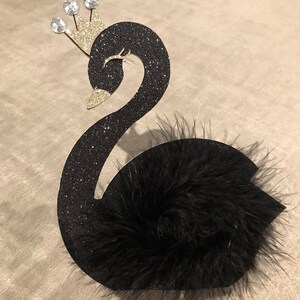 K&ouml;nnte beinhalten: Schwarze Glitzer-Schwan-Kuchenaufsatz mit einer goldenen Krone, die mit drei klaren Edelsteinen verziert ist. Der Schwan hat einen flauschigen schwarzen Feder-Schwanz und ist an einem Holzstab befestigt.