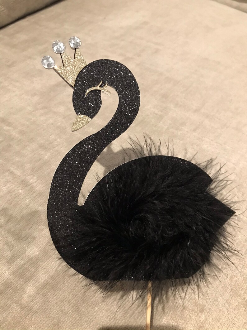 K&ouml;nnte beinhalten: Schwarze Glitzer-Schwan-Kuchenaufsatz mit einer goldenen Krone, die mit drei klaren Edelsteinen verziert ist. Der Schwan hat einen flauschigen schwarzen Feder-Schwanz und ist an einem Holzstab befestigt.