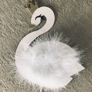K&ouml;nnte beinhalten: Ein wei&szlig;er Schwan-Cake-Topper mit goldener Krone und flauschigen wei&szlig;en Federn. Der Schwan hat ein schwarzes Auge und einen goldenen Schnabel. Der Topper ist an einem Holzstab befestigt.