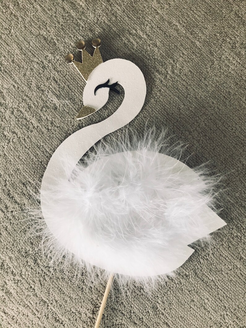 K&ouml;nnte beinhalten: Ein wei&szlig;er Schwan-Cake-Topper mit goldener Krone und flauschigen wei&szlig;en Federn. Der Schwan hat ein schwarzes Auge und einen goldenen Schnabel. Der Topper ist an einem Holzstab befestigt.