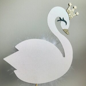 K&ouml;nnte beinhalten: Wei&szlig;er Schwan-Kuchenaufsatz mit goldener Glitzerkrone und Schnabel sowie wei&szlig;en Federn. Der Schwan ist an einem Holzstab befestigt. Der Hintergrund ist einfarbig hellgrau.