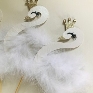 K&ouml;nnte beinhalten: Zwei wei&szlig;e Schwan-Dekorationen mit goldenen Glitzerkronen und flauschigen wei&szlig;en Federakzenten. Die Schw&auml;ne haben schwarze Augen und Schn&auml;bel und sind an Holzst&auml;ben befestigt, ideal f&uuml;r Tortenaufs&auml;tze oder Partydekorationen.