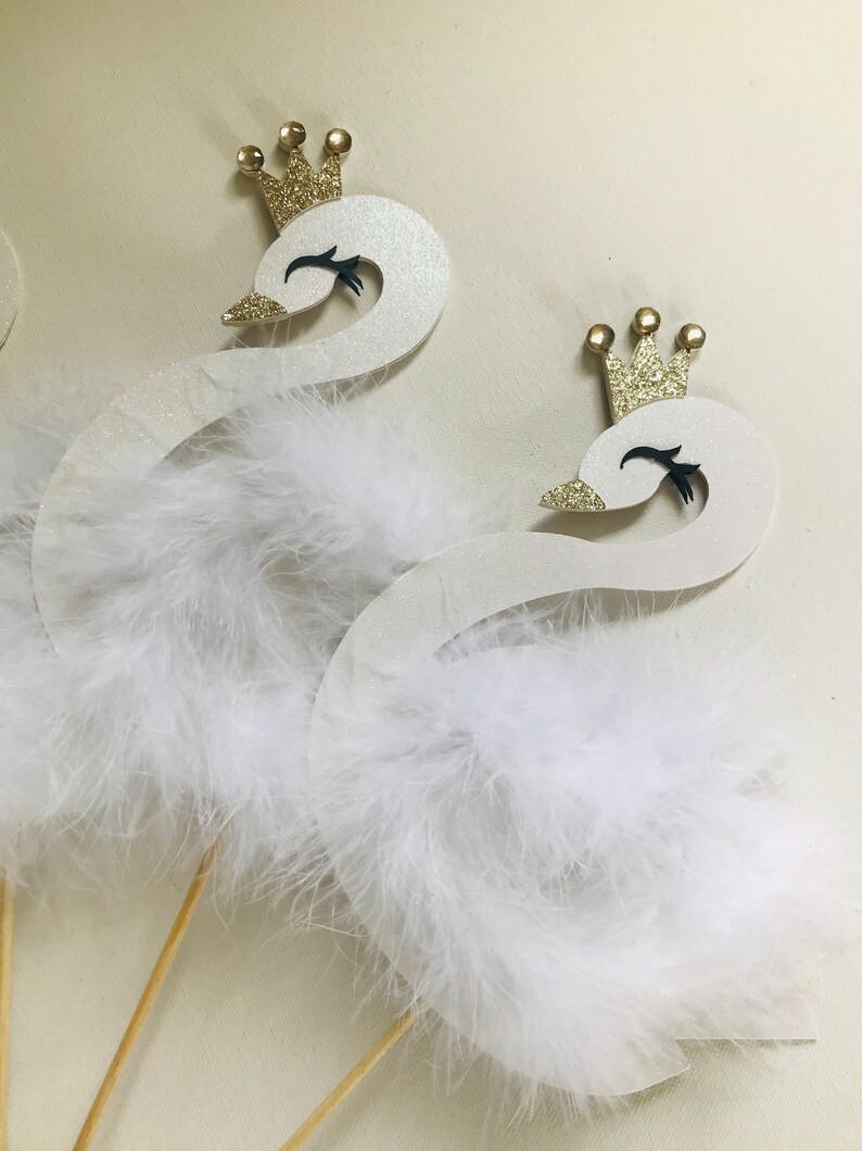 K&ouml;nnte beinhalten: Zwei wei&szlig;e Schwan-Dekorationen mit goldenen Glitzerkronen und flauschigen wei&szlig;en Federakzenten. Die Schw&auml;ne haben schwarze Augen und Schn&auml;bel und sind an Holzst&auml;ben befestigt, ideal f&uuml;r Tortenaufs&auml;tze oder Partydekorationen.