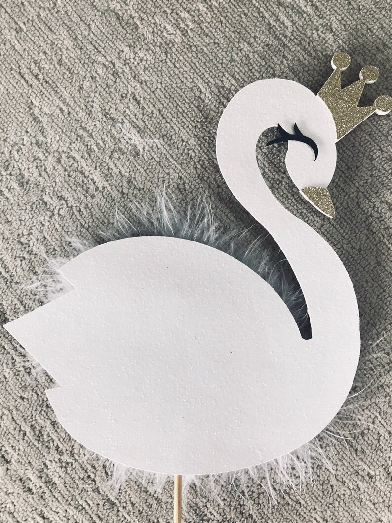 K&ouml;nnte beinhalten: Eine wei&szlig;e schwanf&ouml;rmige Dekoration mit einer goldenen Glitzerkrone und schwarzen Augendetails. Der Schwan hat einen flauschigen wei&szlig;en Federakzent und ist an einem Holzstab befestigt. Dieser Dekorationsartikel ist wahrscheinlich f&uuml;r eine Party oder Veranstaltung.