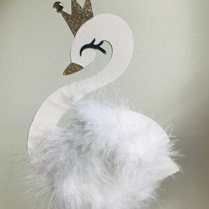 K&ouml;nnte beinhalten: Eine wei&szlig;e schwanf&ouml;rmige Dekoration mit einer goldenen Glitzerkrone und drei goldenen Edelsteinen. Der Schwan hat einen flauschigen wei&szlig;en Federk&ouml;rper und ein schwarzes Auge. Die Dekoration ist auf einem d&uuml;nnen Holzstab befestigt.