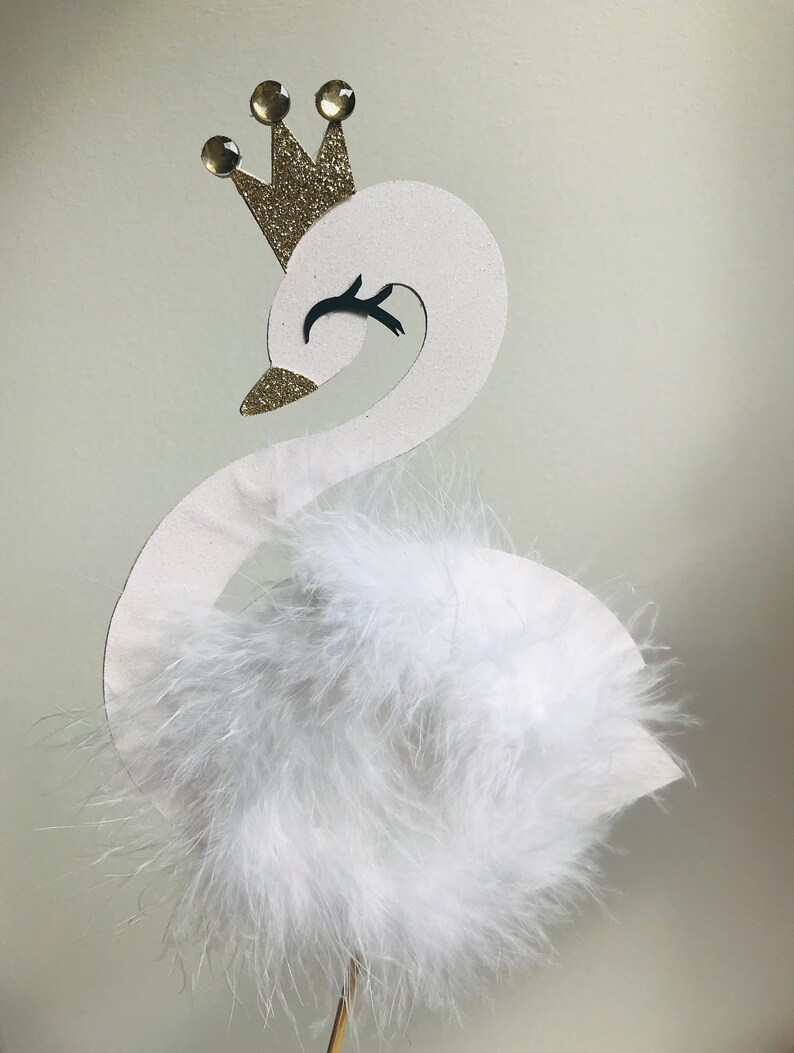 K&ouml;nnte beinhalten: Eine wei&szlig;e schwanf&ouml;rmige Dekoration mit einer goldenen Glitzerkrone und drei goldenen Edelsteinen. Der Schwan hat einen flauschigen wei&szlig;en Federk&ouml;rper und ein schwarzes Auge. Die Dekoration ist auf einem d&uuml;nnen Holzstab befestigt.