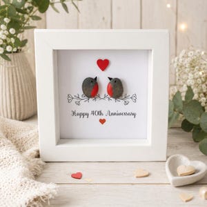 May include: A square white frame containing pebble art of two birds with red chests perched on a black line drawing of branches. Above the birds is a red heart, and below them is the text Happy 40th Anniversary with a small red heart underneath.