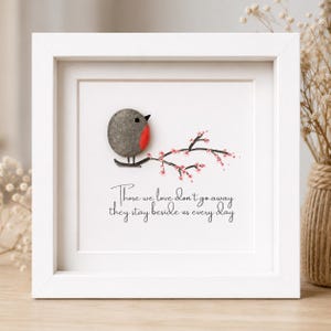 May include: A square white frame containing pebble art of a robin perched on a branch with pink blossoms. The text reads: Those we love don't go away, they stay beside us every day. The bird is made of a grey stone with a painted red breast.